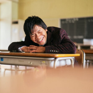 教室の机に腕を組んで顔をのせ、思わず笑い出すタキシード姿の新郎。学校ロケの前撮りで撮れた自然な笑顔