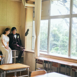 大きな窓から緑が見える教室で、窓際に腰を下ろして寄り添う新郎新婦。懐かしい机と椅子が並ぶ学校ロケーションフォトの一枚