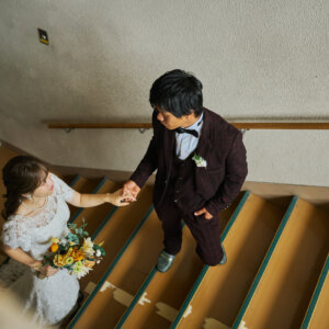 校舎の階段を上から見下ろした構図で、ひまわりのブーケを持つ花嫁の手を引く新郎。学校で撮る洋装前撮りならではのアングル