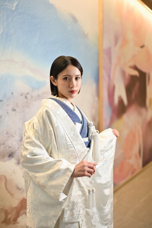 青い襟元をのぞかせた白無垢を着て、鶴の絵のアートウォールの前に立つ花嫁。和装前撮りで選べる白無垢AS103の衣装カタログ写真