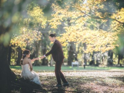 黄葉した大木の下で、座る花嫁に手を差し伸べるタキシードの新郎。砧公園の木漏れ日の中で撮った秋のウェディングドレス前撮り
