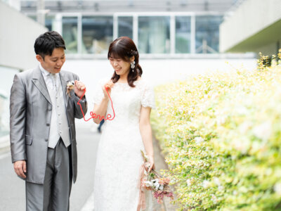 グリーンの植え込みのそばで、赤い糸を小指に結んでほほえむ新郎新婦。南青山の街で撮る洋装前撮り
