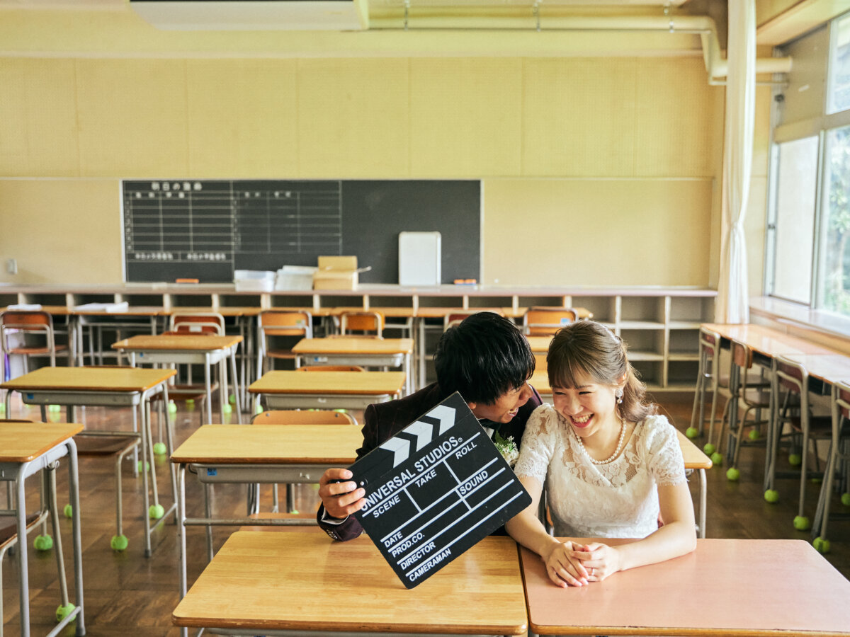 教室の机に並んで座り、カチンコを手に笑う新郎新婦。映画のワンシーンのような演出で撮る学校の前撮り