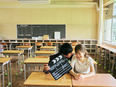 教室の机に並んで座り、カチンコを手に笑う新郎新婦。映画のワンシーンのような演出で撮る学校の前撮り