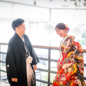 窓から光が入る階段の吹き抜けで、笑い合う紋付袴の新郎と赤い色打掛の花嫁。明るい和装スタジオフォト