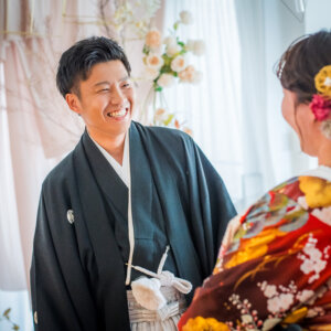 花で飾られた明るいスタジオで、髪飾りを付けた色打掛の花嫁を見つめて笑う紋付袴の新郎。和装フォトウェディングの自然な表情
