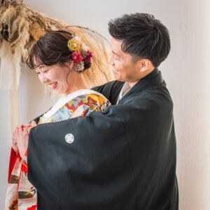 後ろから花嫁を抱きしめて笑う紋付袴の新郎と、照れてうつむく鶴柄の色打掛の花嫁。ハグのポーズで撮る和装スタジオフォト