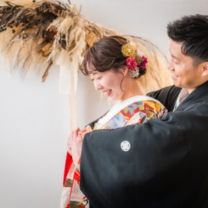 パンパスグラスを飾った白壁の前で、色打掛の花嫁を後ろから包むように抱きしめる紋付袴の新郎。南青山のスタジオで撮る和装前撮り