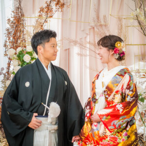 花で飾られたセットの前で向かい合い、笑顔で見つめ合う紋付袴の新郎と赤い色打掛の花嫁。南青山で撮る和装スタジオフォト