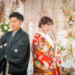 ドライフラワーとバラのセットの前で、腕を組む紋付袴の新郎と目を伏せてほほえむ色打掛の花嫁。クールな和装前撮り