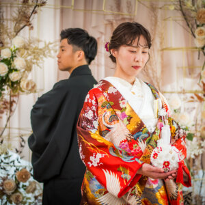 花で飾られたセットで背中合わせに立つ紋付袴の新郎と、ブーケを手に目を伏せる赤い色打掛の花嫁。南青山のスタジオで撮る和装フォトウェディング