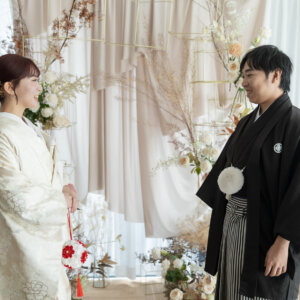 白いカーテンと花のアーチの前で、照れたように笑い合う紋付袴の新郎と色打掛の花嫁。南青山のスタジオフォトで撮る和装前撮り