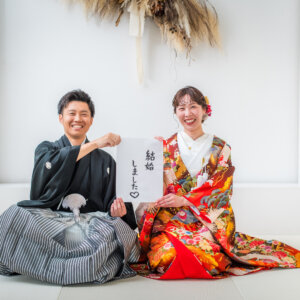 '結婚しました'の手書きボードを持って白い畳に座る紋付袴の新郎と赤い色打掛の花嫁。結婚報告に使える和装スタジオフォト