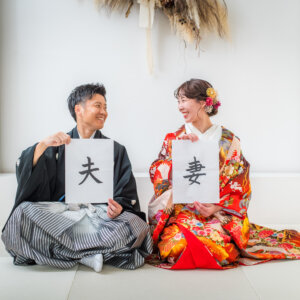 '夫''妻'と書いた紙を持ち、白い畳に座って見つめ合う紋付袴の新郎と色打掛の花嫁。結婚報告にも使える和装前撮り
