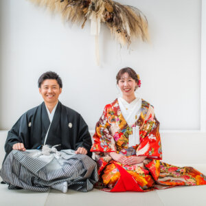 白い畳にあぐらで座って笑う紋付袴の新郎と、ひざに手を重ねる赤い色打掛の花嫁。南青山のスタジオで撮る和装前撮り