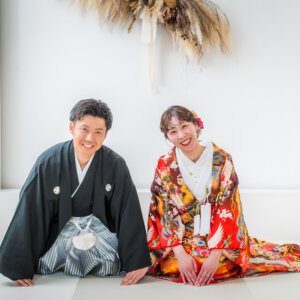 白い畳に両手をついてにっこり笑う紋付袴の新郎と、裾を広げた赤い色打掛の花嫁。白を基調にした和装スタジオフォト