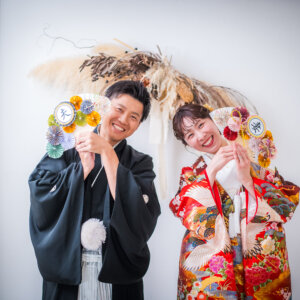 '夫''妻'の扇子を顔の横に掲げて笑う紋付袴の新郎と鶴柄の色打掛の花嫁。小物を使った和装スタジオフォト