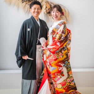 ドライフラワーを飾った白壁の前で寄り添う、鶴柄の赤い色打掛の花嫁と紋付袴の新郎。南青山のスタジオで撮る和装前撮り