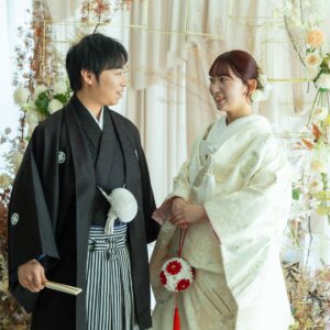 ドライフラワーとバラの装飾の前で向かい合いほほえむ紋付袴の新郎と色打掛の花嫁。南青山のスタジオで撮るナチュラルな和装前撮り