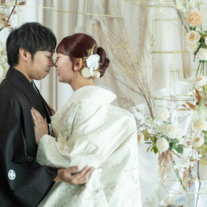 花の装飾に囲まれておでこを寄せ合い抱き合う黒紋付袴の新郎と白地の色打掛の花嫁。スタジオで撮る和装前撮りのおでこポーズ