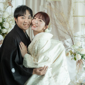 白いバラの装飾の前で、頬を寄せて笑顔で抱き合う紋付袴の新郎と色打掛の花嫁。南青山のスタジオフォトで撮る仲良しな結婚写真