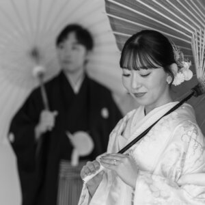 和傘をさしてうつむく色打掛の花嫁と、後ろに立つ紋付袴の新郎をモノクロで写した一枚。スタジオで撮る和装前撮りのしっとりカット