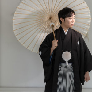 白い和傘を肩にかけて遠くを見つめる黒紋付袴の新郎。南青山のスタジオで撮る和装前撮りの新郎ソロカット