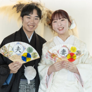 '夫''妻'と書かれた扇子を持って笑顔で並ぶ黒紋付袴の新郎と色打掛の花嫁。スタジオで撮る和装前撮りの人気アイテム