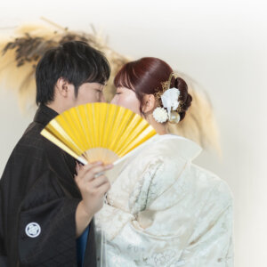 金色の扇子で口元を隠してキスをする紋付袴の新郎と白地の色打掛の花嫁。南青山のスタジオで撮る和装前撮りの扇子ポーズ
