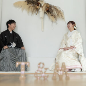 イニシャルのオブジェ越しに、座って笑い合う黒紋付袴の新郎と白地の色打掛の花嫁。南青山のスタジオで撮る和装の結婚写真