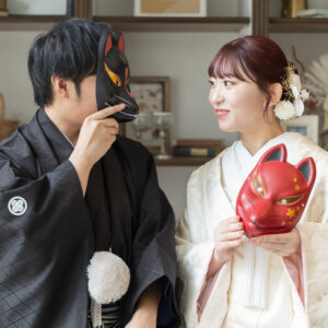 黒と赤の狐のお面を手に寄り添う紋付袴の新郎と白地の色打掛の花嫁。南青山のスタジオで撮る和装前撮りのユニークなアイテム