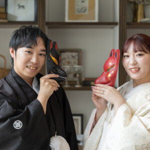 狐のお面を顔の横に持って笑う黒紋付袴の新郎と色打掛の花嫁。本棚のあるスタジオで撮る遊び心のある和装前撮り
