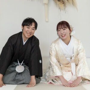 白い床に並んで座り、手をついて笑顔を見せる紋付袴の新郎と白地の色打掛の花嫁。南青山のスタジオで撮るかわいい和装前撮り