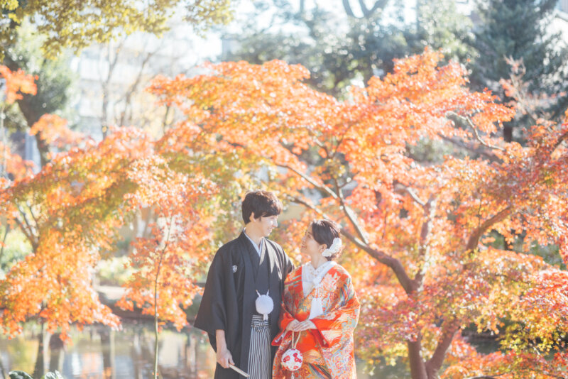 鮮やかなオレンジ色の紅葉と池を背に、見つめ合う紋付袴の新郎と赤い色打掛の花嫁。秋の和装前撮り