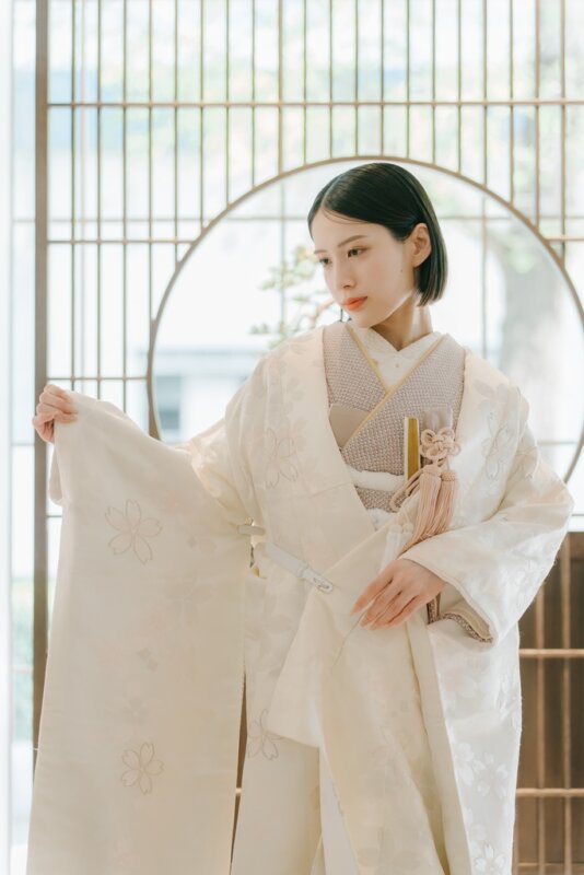 丸窓と格子戸のある和室で袖を広げ、桜の刺繍を見せる白無垢の花嫁。柔らかな光が差す和装前撮りの白無垢AS102