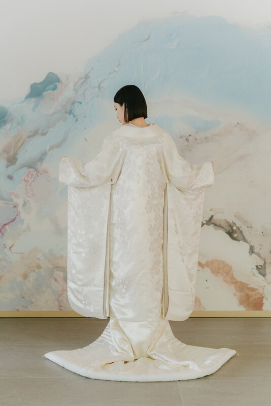 淡いブルーの壁画の前で両袖を広げ、後ろ姿を見せる白無垢の花嫁。背中から裾まで続く地紋が分かる和装フォトウェディングのバックスタイル