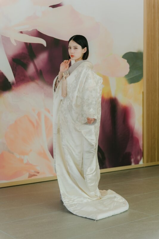 花柄の壁を背景に、筥迫の房を手に横向きで立つ白無垢姿の花嫁。裾を引いた全身が美しいスタジオフォトの白無垢AS102