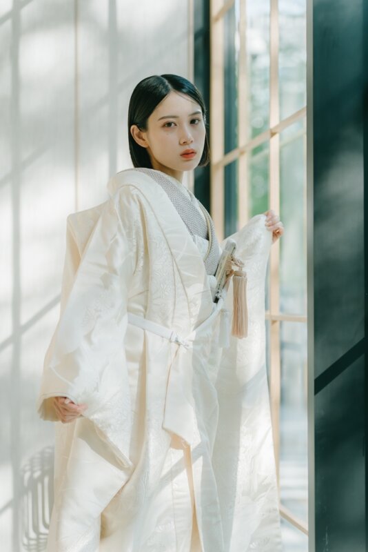 格子窓から光が差し込む廊下で、袖を広げてこちらを見つめる白無垢の花嫁。ピンクの房飾りが映える白無垢の和装前撮り