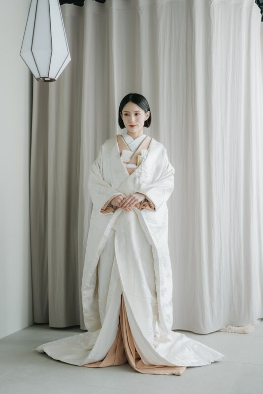 ペンダントライトが下がるスタジオで両手を重ねて立つ、裾を広げた白無垢の花嫁。正面から撮った和装の結婚写真