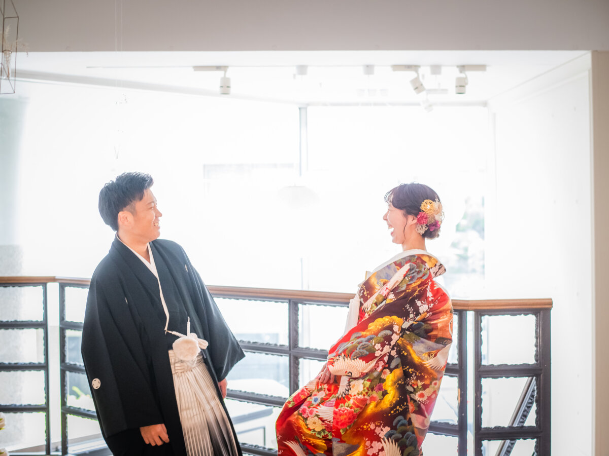 光が差しこむ窓辺の手すりの前で、向かい合って笑う紋付袴の新郎と赤い色打掛の花嫁。南青山のスタジオで撮る和装フォトウェディング